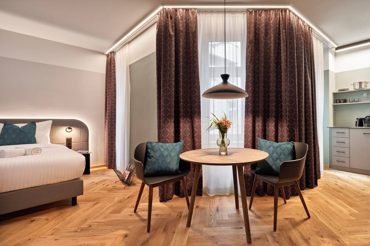Ein modernes Hotelzimmer mit Bett, Esstisch mit Stühlen, einer Küchenzeile und einem großen Fenster mit Vorhängen.
