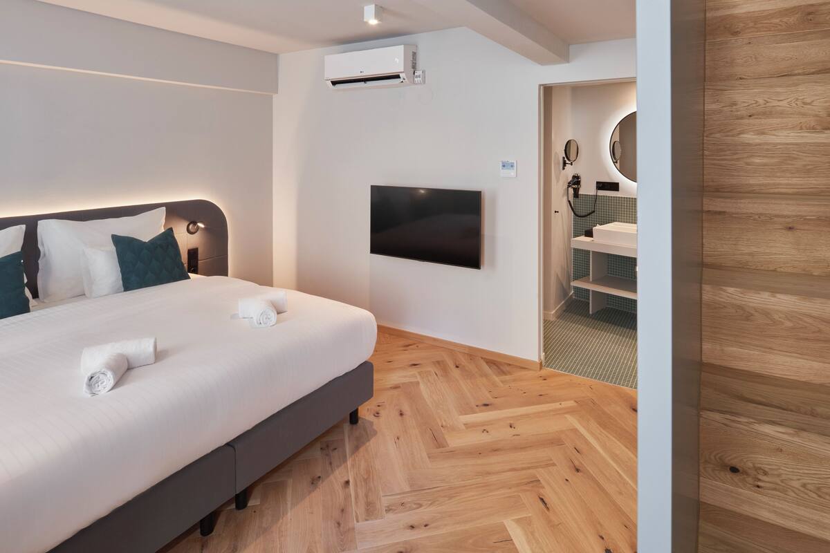 Ein modernes Hotelzimmer mit einem Bett, einem an der Wand befestigten Fernseher und einem Badezimmer mit Spiegel und Waschbecken.