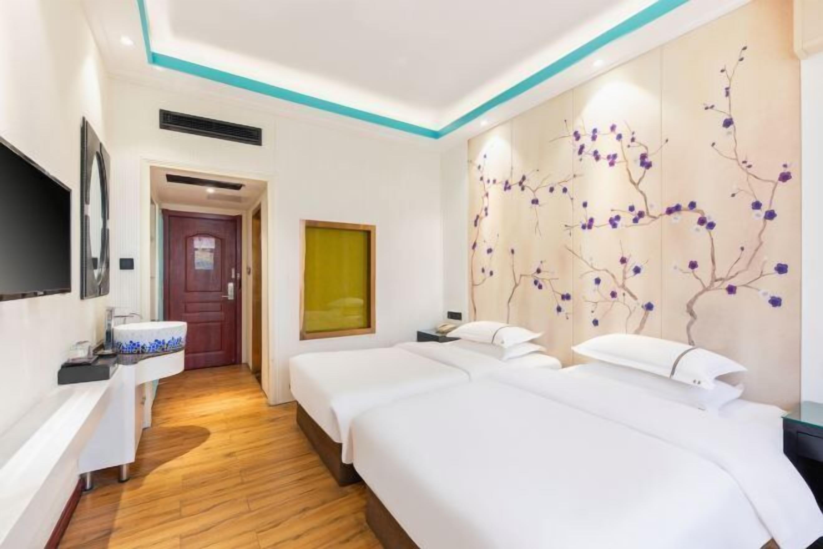 A hotel room with two beds, a TV, a bathroom with a sink, and a decorative wall with branches.