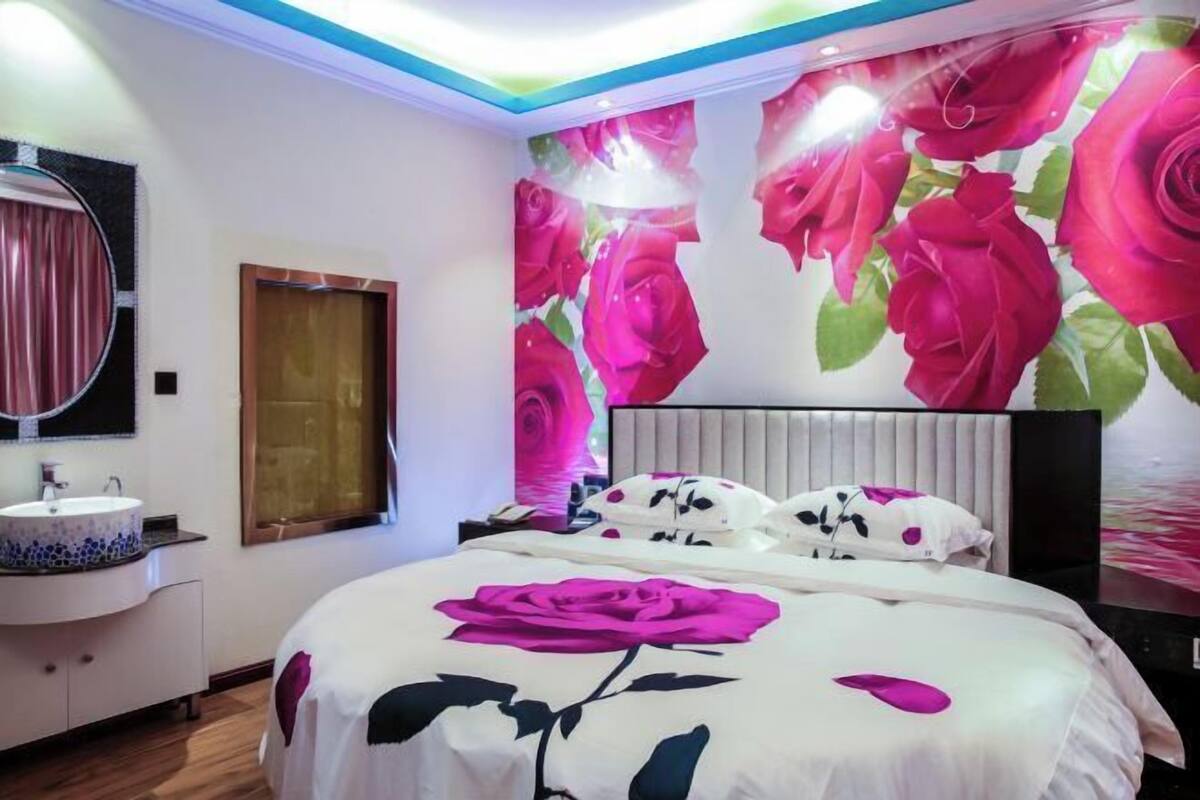 Een slaapkamer met een bed, nachtkastje, spiegel, ronde spiegel, raam en wand met bloemenmotief.