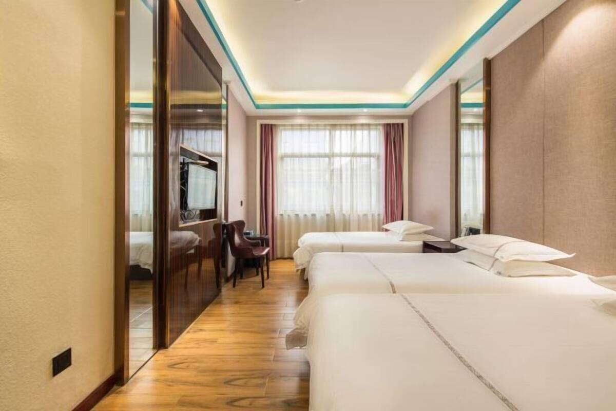 Hotelkamer met houten vloer, bureau met stoel, twee bedden en een groot raam met gordijnen.