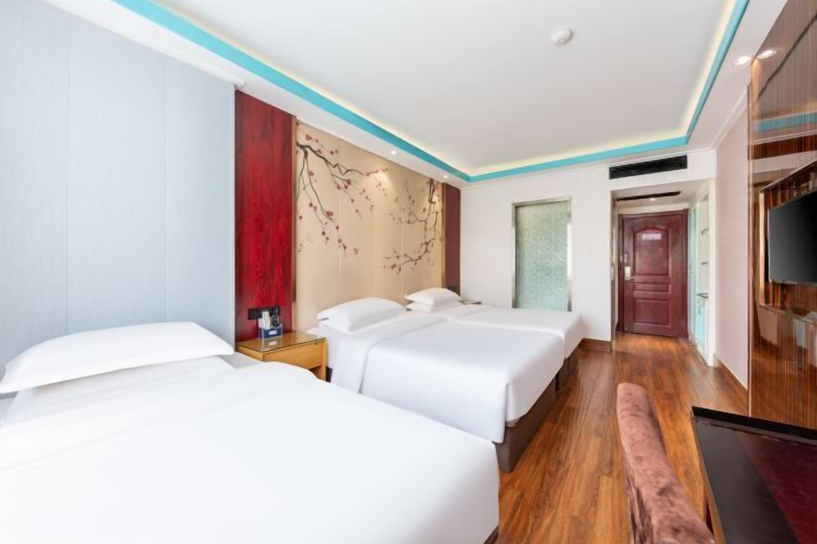 A hotel room with two beds, a wooden floor, a wall with a decorative panel, and a television.