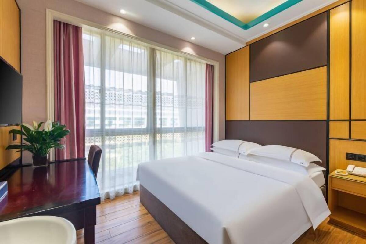 Een hotelkamer met een groot bed, een bureau met stoel, een kamerplant en een raam met gordijnen.