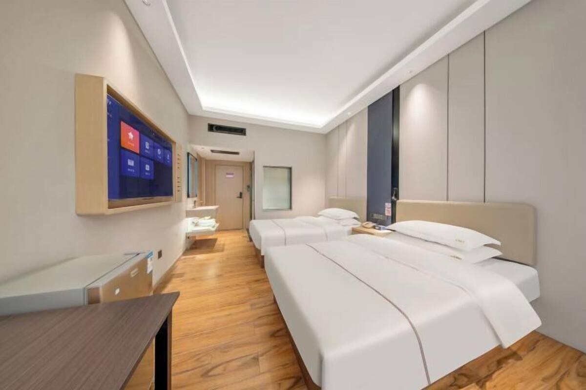 Een hotelkamer met een houten vloer, een bed met witte beddengoed, een bureau, een aan de muur bevestigde televisie en een deur die naar een andere kamer leidt.