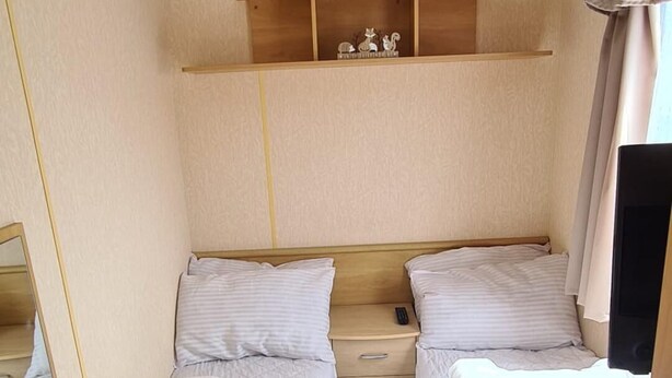 A compact room with two single beds, a small shelf with decorative items, a ceiling light, and a window with curtains. - 8 Birth Caravan Based on Coral Beach, Ingoldmells (Skegness)