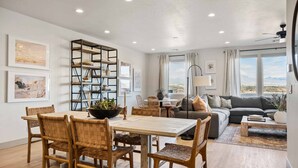A spacious living room with a dining area, a sofa, and a coffee table. There are framed pictures on the walls and a large window with curtains. - Casa Lua: Desert Color Lagoon + Beaches (St. George)