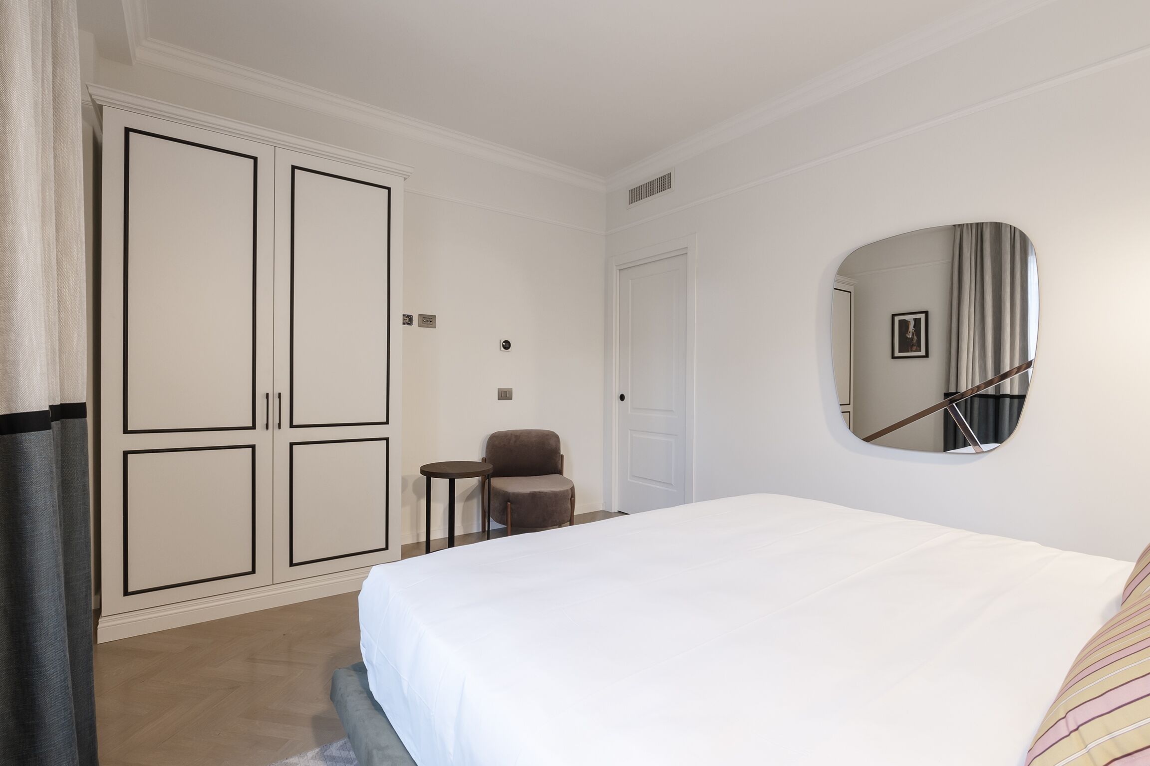 A bedroom with a large bed, a wardrobe, a chair, and a mirror on the wall.