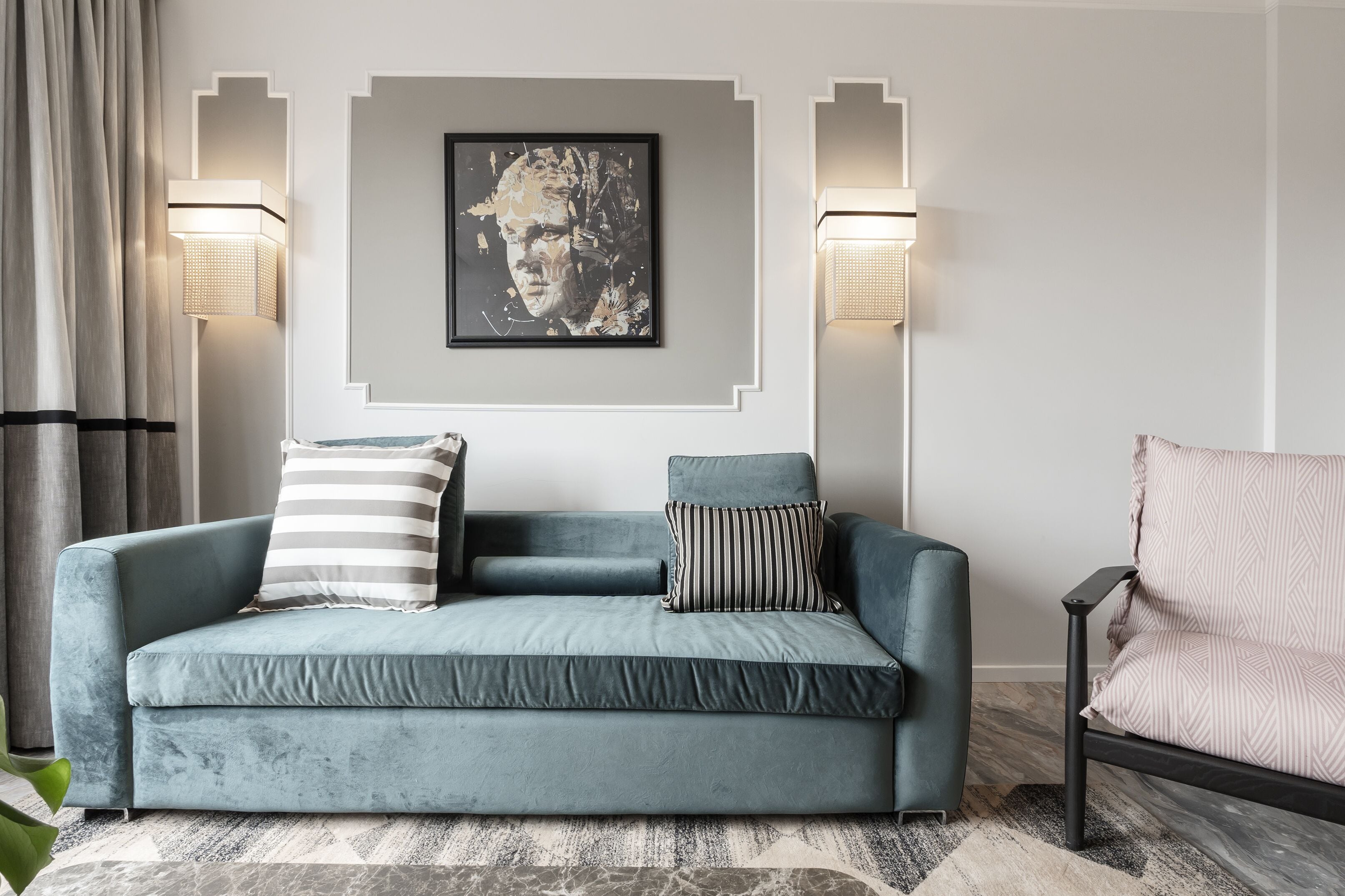 Uma sala de estar moderna com um sofá azul-turquesa, almofadas listradas e estampadas, uma obra de arte emoldurada acima e duas luminárias fixadas na parede.