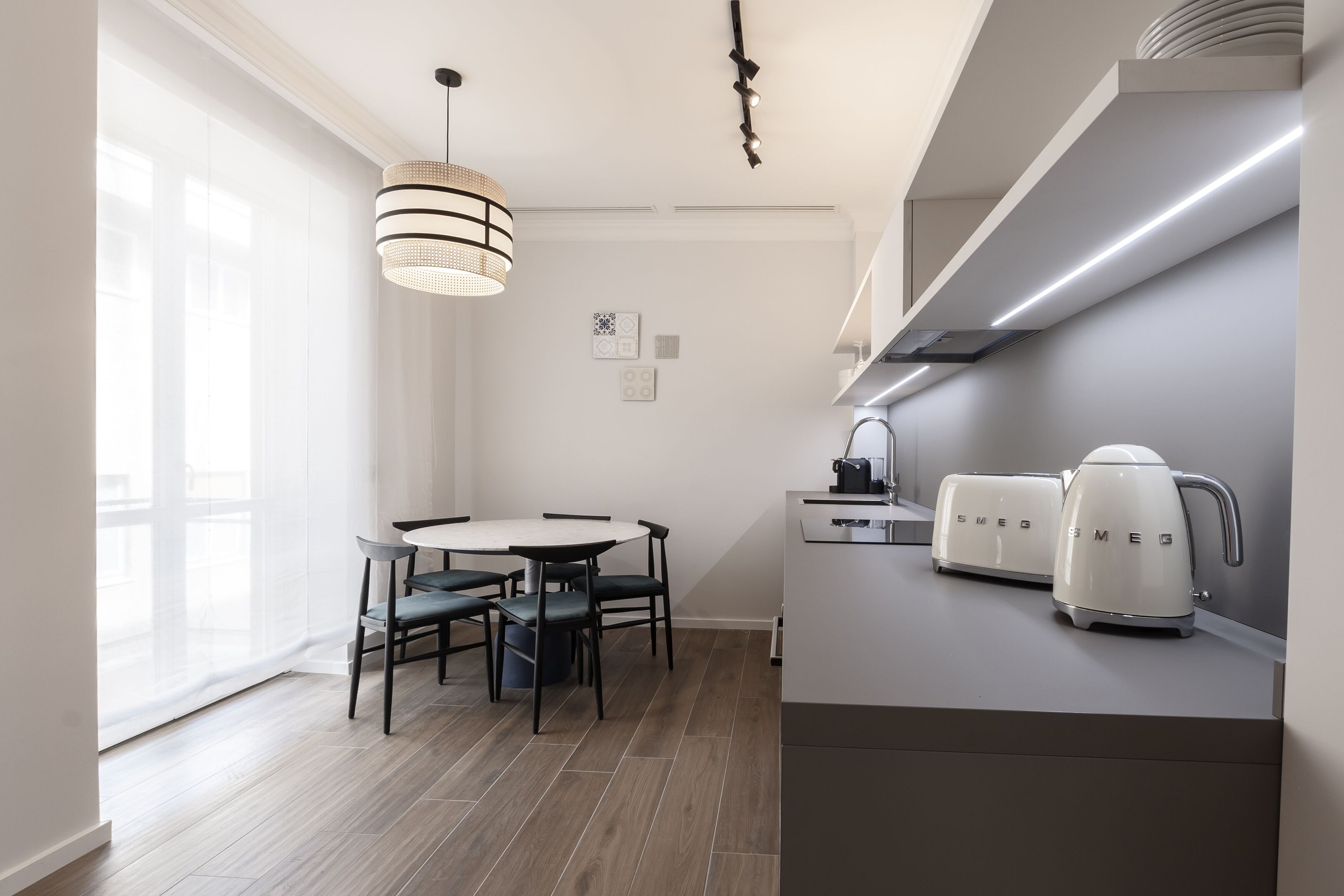 Une cuisine moderne avec une table de salle à manger ronde, deux chaises, un luminaire suspendu et deux bouilloires électriques blanches posées sur le plan de travail.