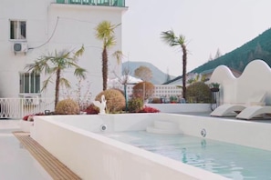 Zona de piscina con muelle de madera, palmeras y un edificio blanco con montañas al fondo.