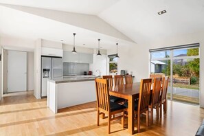 Cocina moderna con una isla central, mesa de comedor de madera y sillas, y vistas a un jardín a través de ventanas amplias.