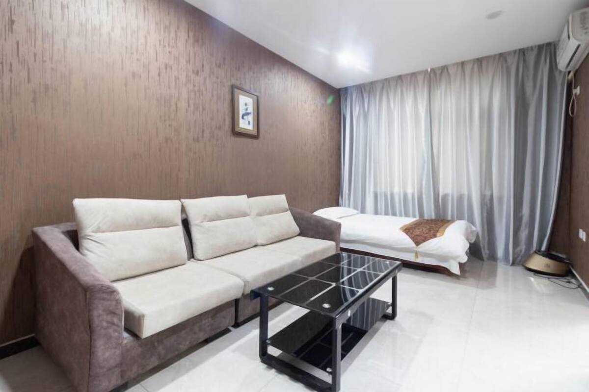 A modern room with a beige sofa, a glass-top coffee table, a bed with white bedding, and a large window with curtains.