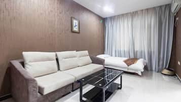 Een moderne kamer met een beige bank, een salontafel met glazen blad, een bed met wit beddengoed en een groot raam met gordijnen.