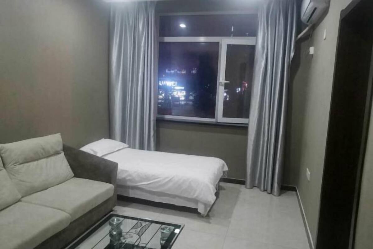 A small, well-lit room with a sofa, a single bed, a glass coffee table, and a window with curtains.