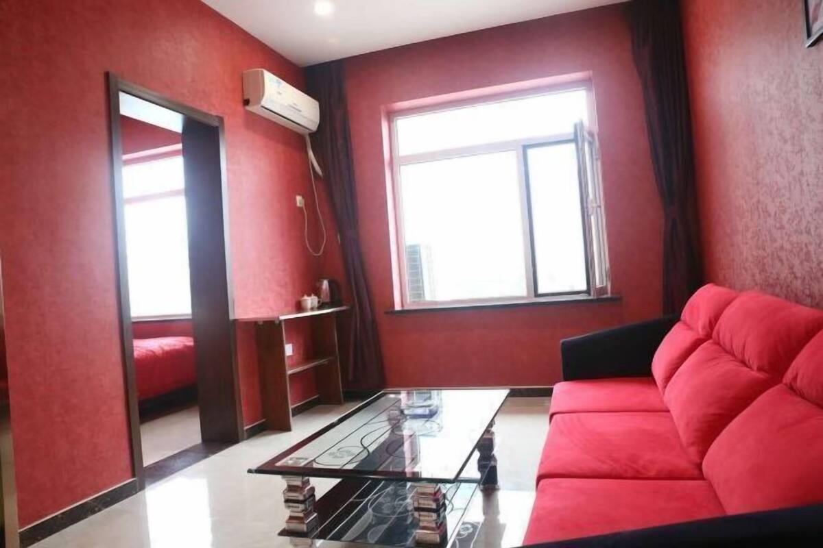 A room with a red sofa, a glass coffee table, a red bed, and a window with curtains.