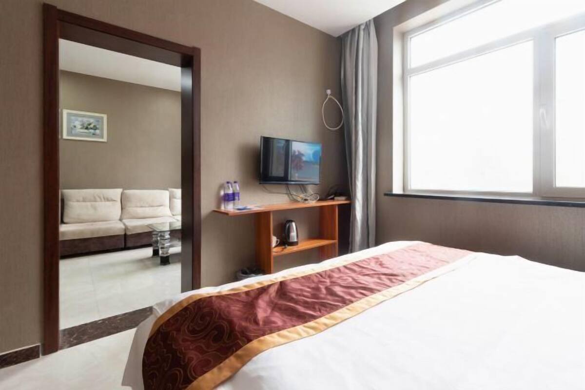 A hotel room with a bed, a television on a stand, a sofa in the background, and a window with curtains.
