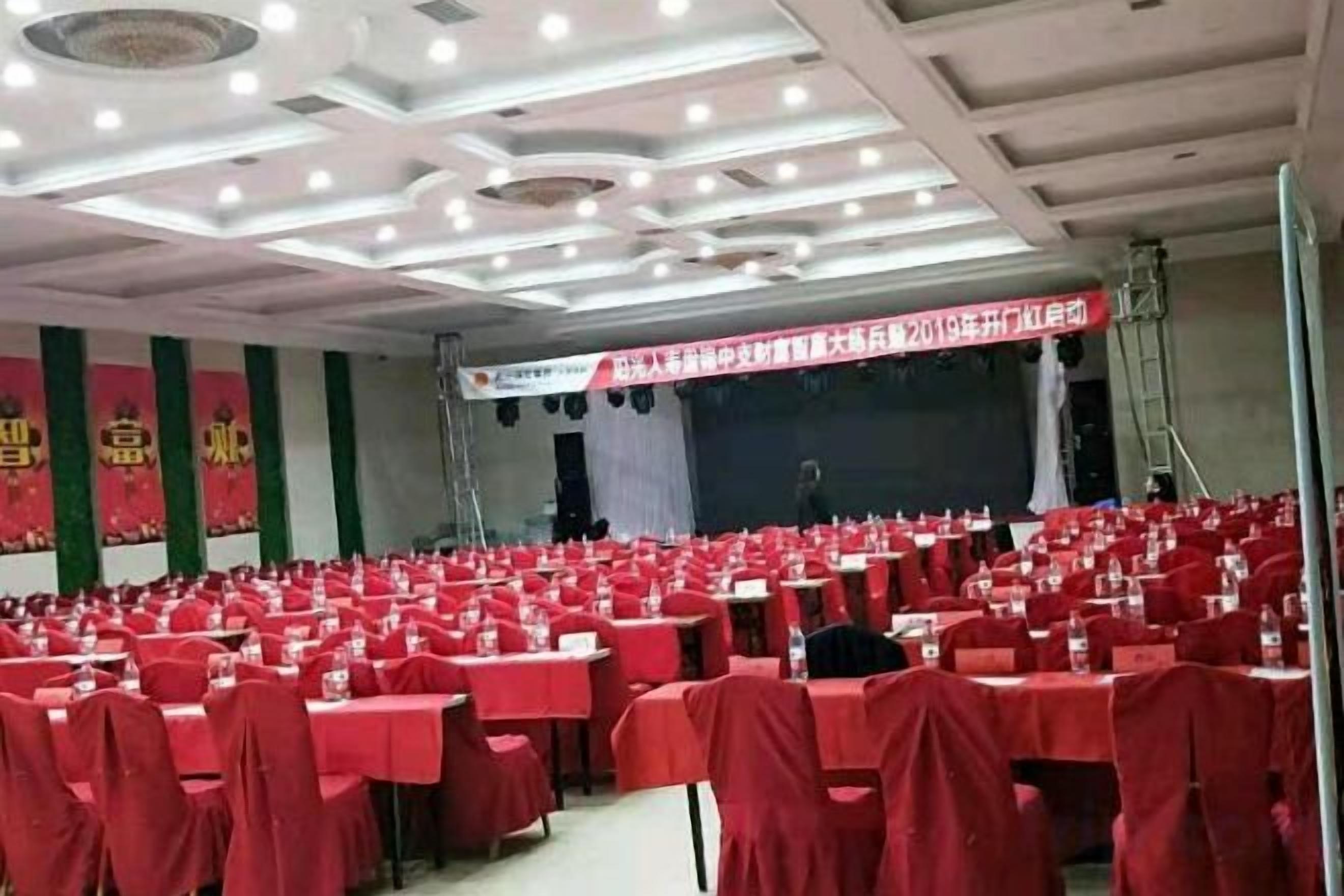 Une salle de conférence avec des rangées de tables et de chaises, une scène ornée d’une banderole et un pupitre pour l’orateur.