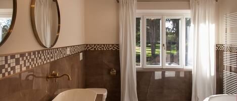 A bathroom with a large oval sink, a toilet, and a bathtub. There is a window with curtains, a chandelier, and a tiled floor.