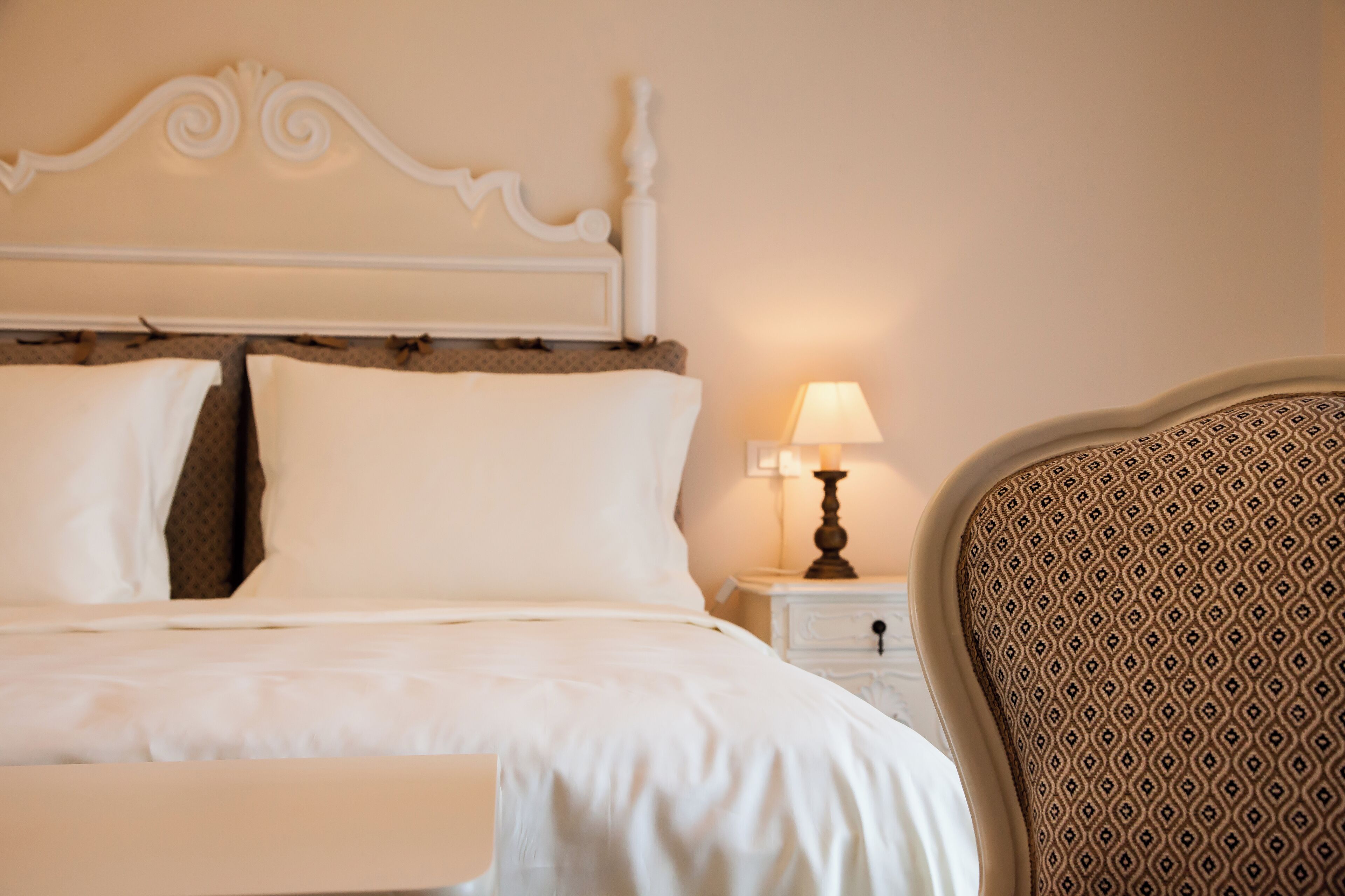 A neatly made bed with white linens, a white headboard, a bedside table with a lamp, and a patterned armchair.