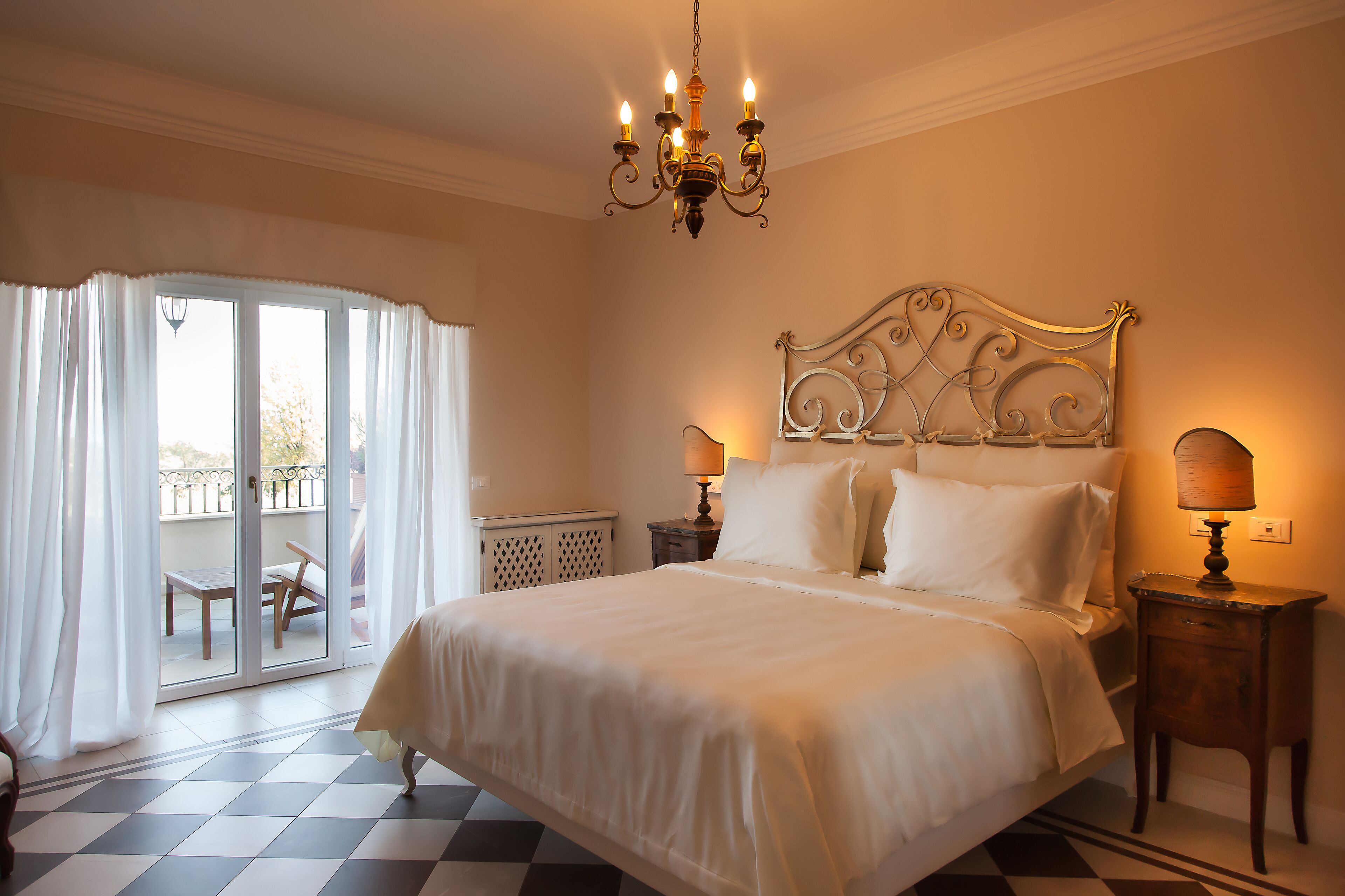 Ein Hotelzimmer mit einem großen Bett, einer Kristallleuchtre, einem Nachttisch und Blick durch eine Glastür in einen Außenbereich.