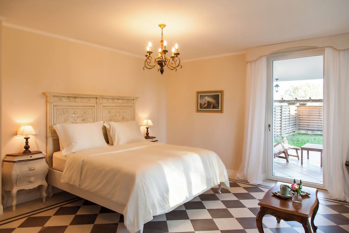 Une chambre à coucher avec un lit, deux tables de chevet avec des lampes, un lustre, un sol à carreaux, une petite table avec un vase et une fleur, et une porte-fenêtre coulissante donnant sur un balcon.