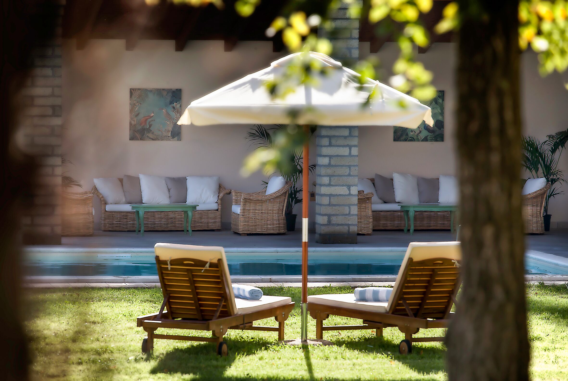 A poolside area with two wooden lounge chairs, a white umbrella, and wicker furniture.