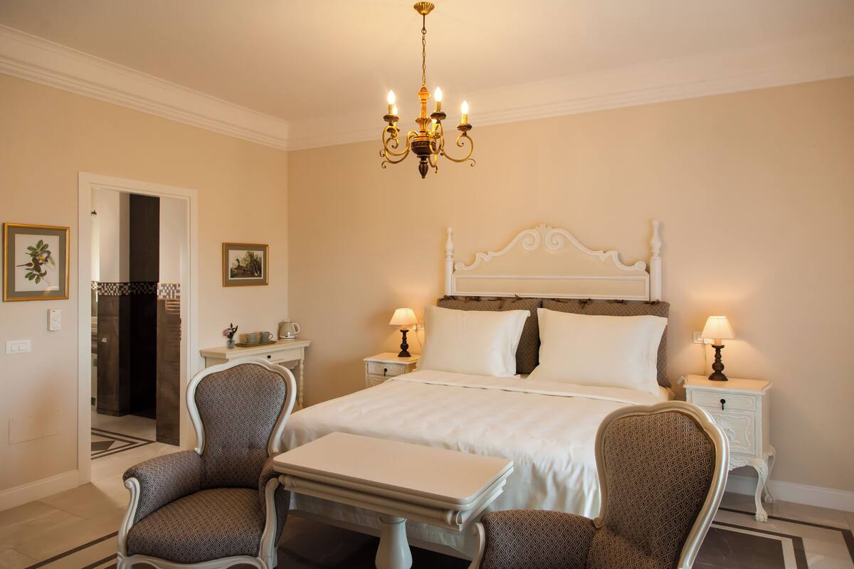 Une chambre d’hôtel avec un grand lit, deux tables de chevet avec des lampes, une petite table, deux chaises à motifs et un lustre.