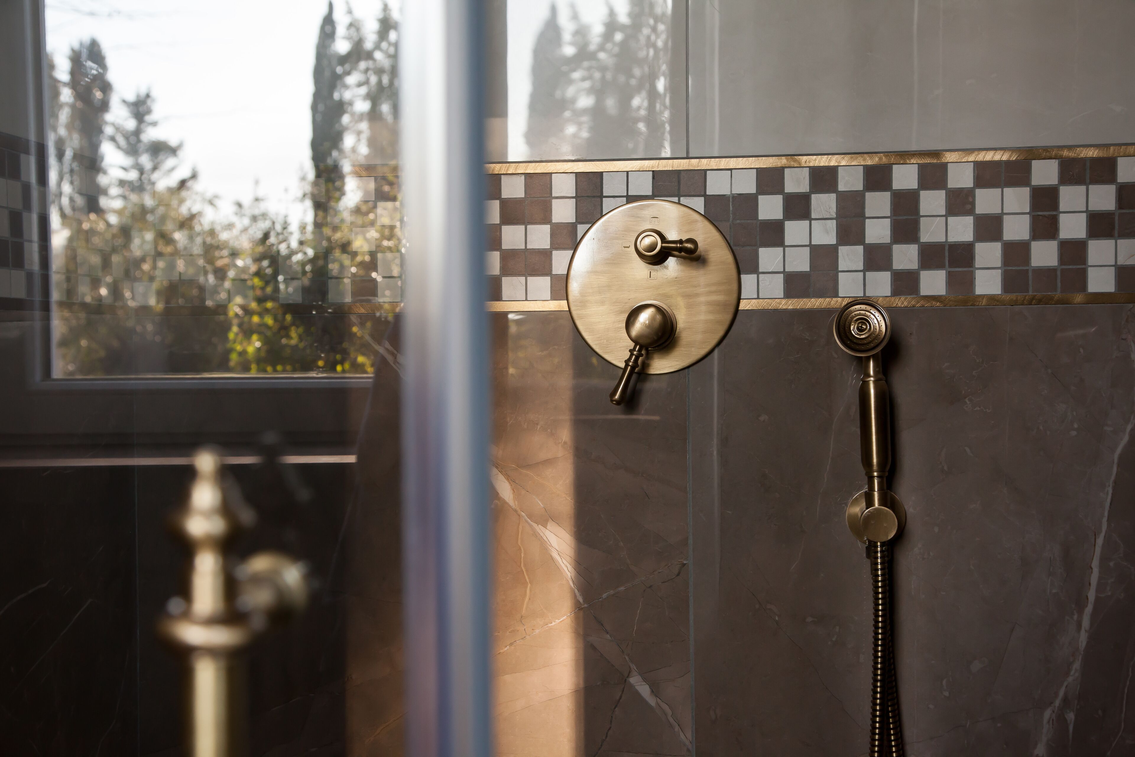 A bathroom with a shower control panel, a handheld showerhead, and a view of trees outside.