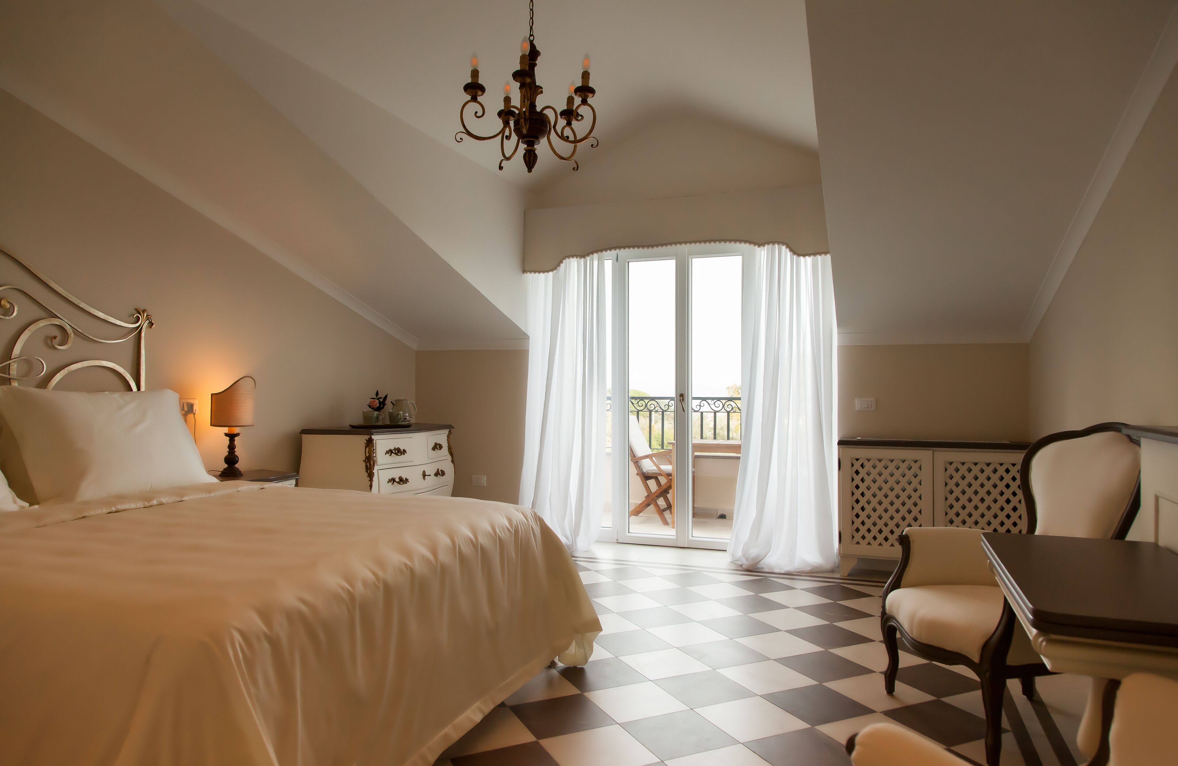 A bedroom with a large bed, a chandelier, a table, and a balcony with curtains.
