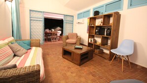 A living room with a sofa, a coffee table, a bookshelf, and a chair. There is a dining area visible through an open door.