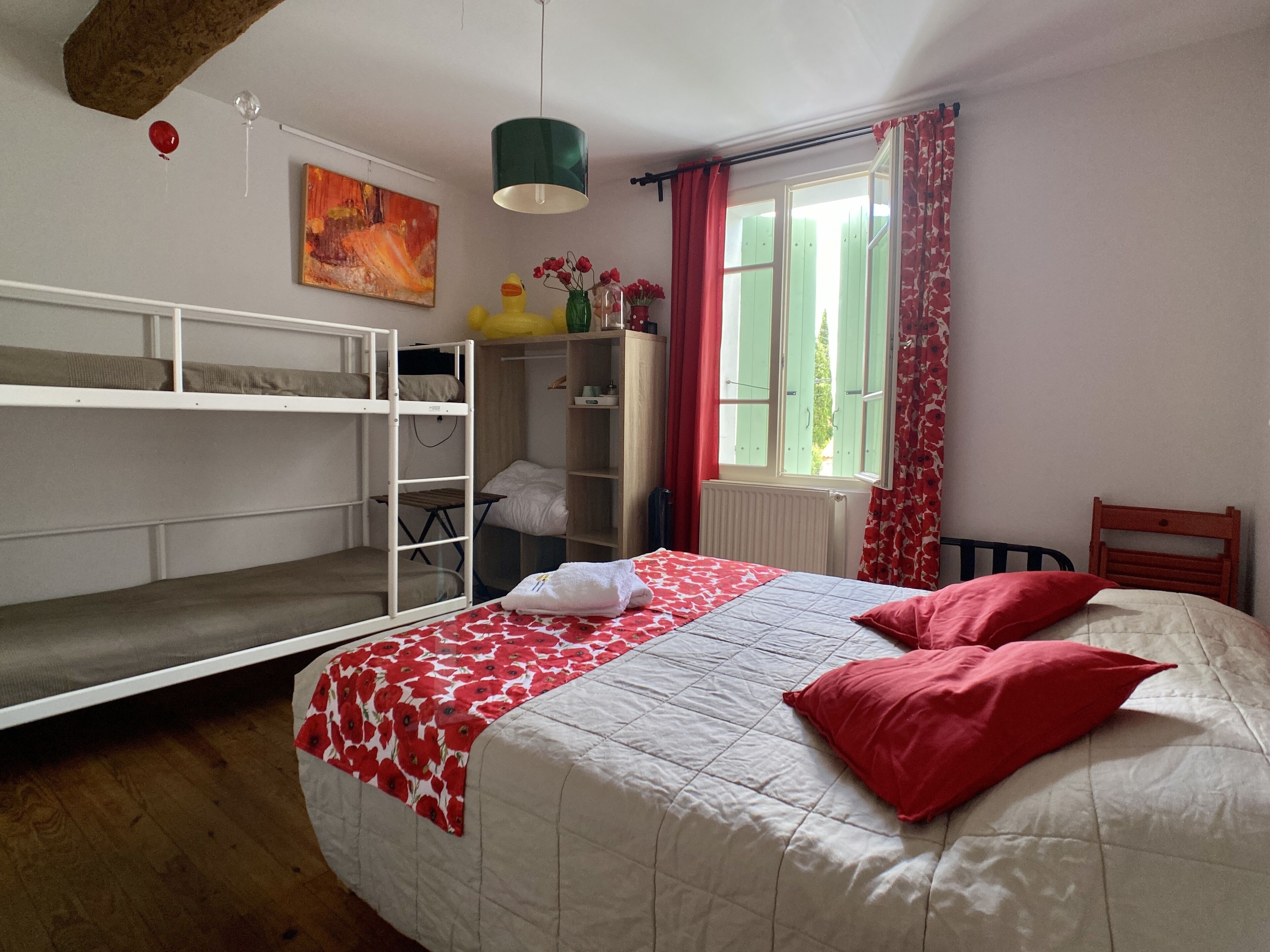 A bedroom with bunk beds, a single bed with red pillows, a window with red curtains, and a green pendant light.