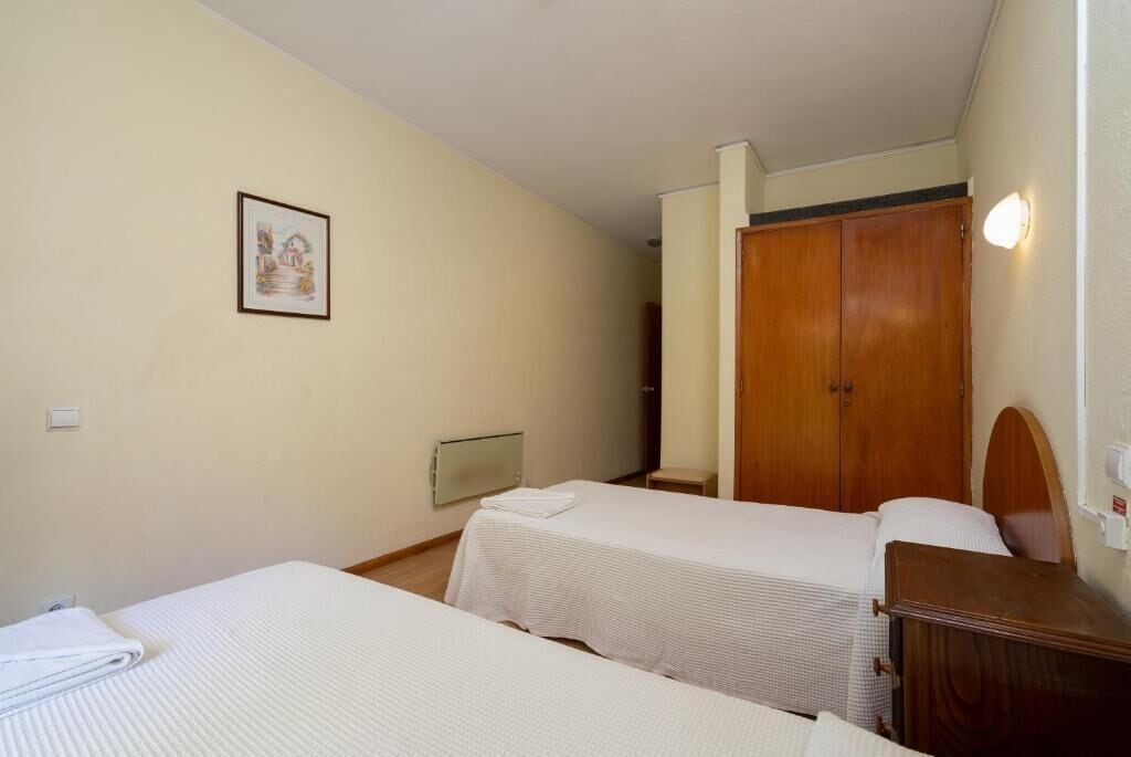 A hotel room with two beds, a wooden wardrobe, a radiator, and a framed picture on the wall.