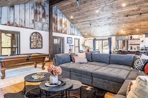 A spacious living room with a pool table, grey sectional sofa, and a rustic wooden ceiling.