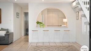 A modern kitchen with white cabinets, a central island with stools, and a staircase with stone steps. - Alice (Bayona 7) - Little Cove (Noosa Heads)