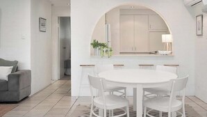 A modern living room with a white dining table, white chairs, a grey sofa, and a kitchen with a large arched window. - Alice (Bayona 7) - Little Cove (Noosa Heads)