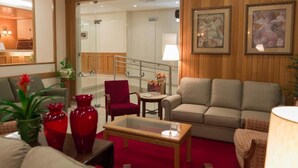 Um saguão de hotel com carpete vermelho, uma poltrona vermelha, um sofá bege e uma mesa de centro de vidro. Há quadros nas paredes e um abajur sobre uma mesa lateral.