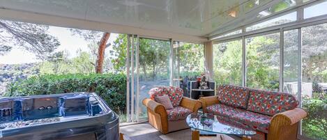 A modern indoor space with a hot tub, wicker furniture, and large windows offering a view of greenery.