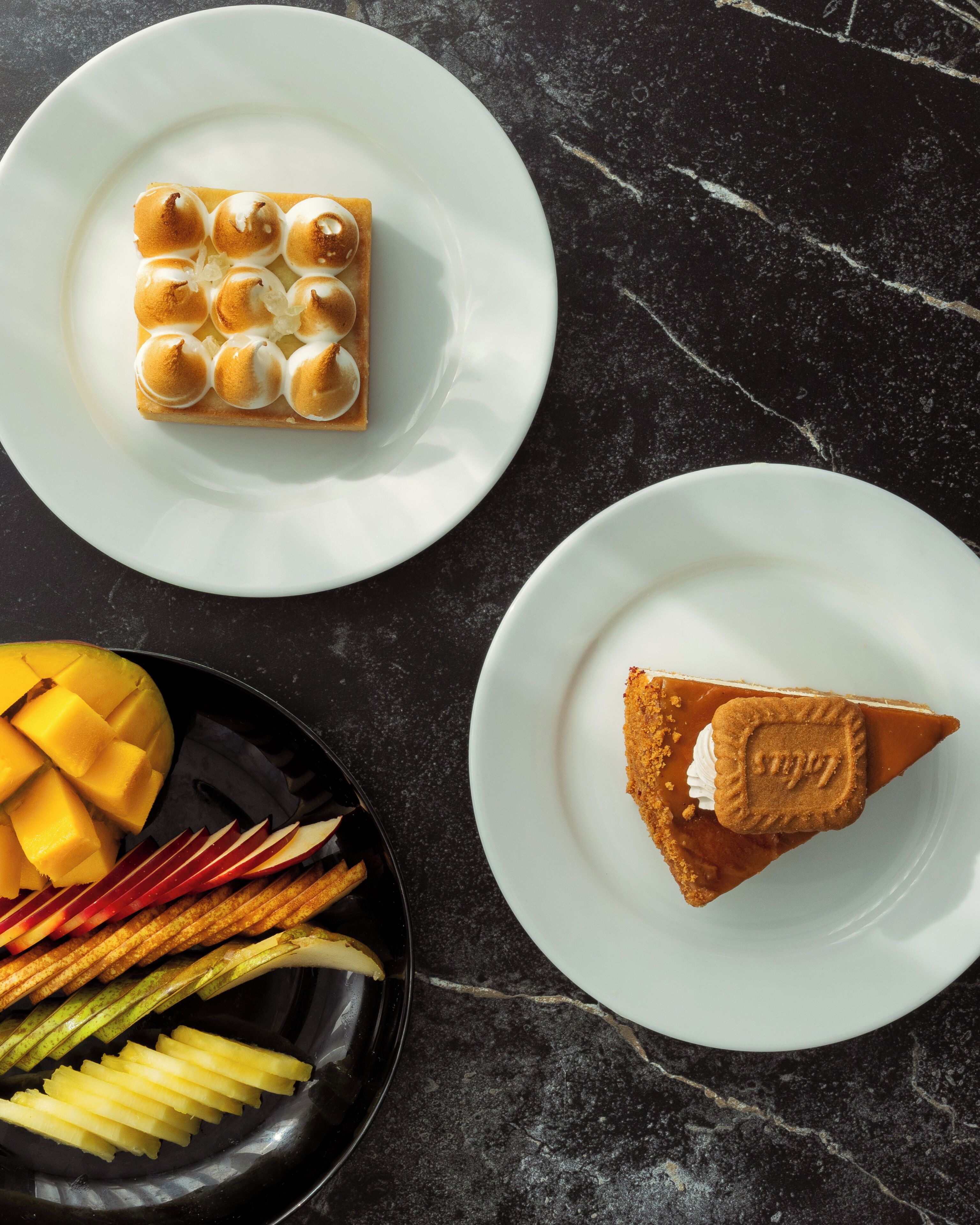 Um prato de sobremesa com um pastel quadrado coberto com merengue, uma fatia de torta com biscoito, e uma tigela de frutas e legumes fatiados.