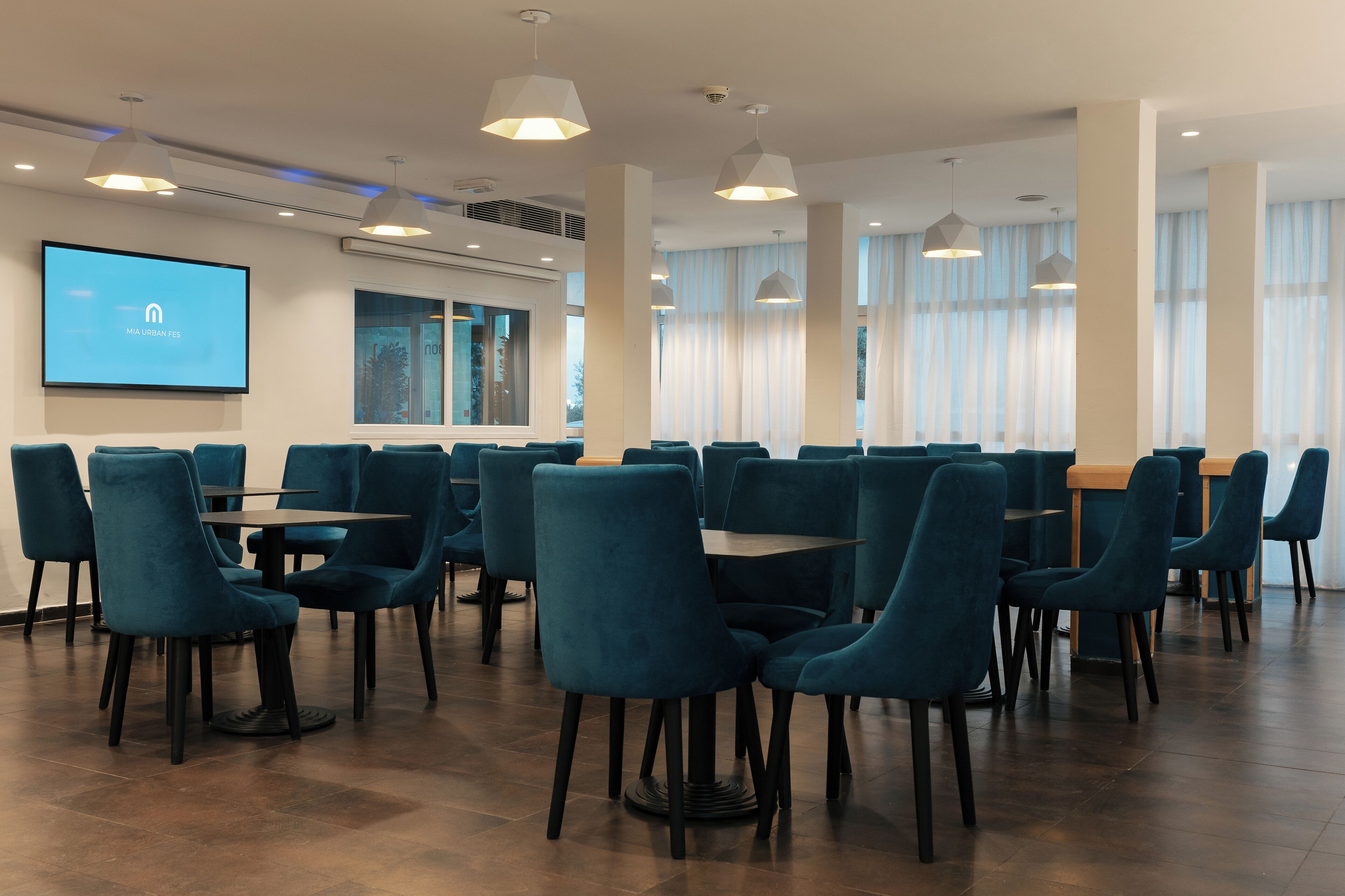 Un salón de conferencias con sillas y mesas color turquesa, ventanas amplias con cortinas y una pantalla mostrando un logotipo.