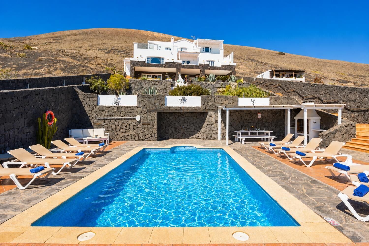 A swimming pool with lounge chairs, a stone wall, and a white building in the background.