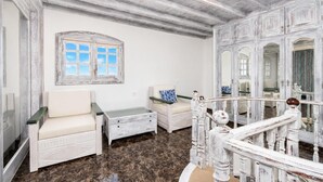 Ein Zimmer mit Marmorboden, einem weißen Sofa, einem kleinen Tisch, einem Fenster mit blauen Vorhängen und einer Treppe mit weißen Geländern.