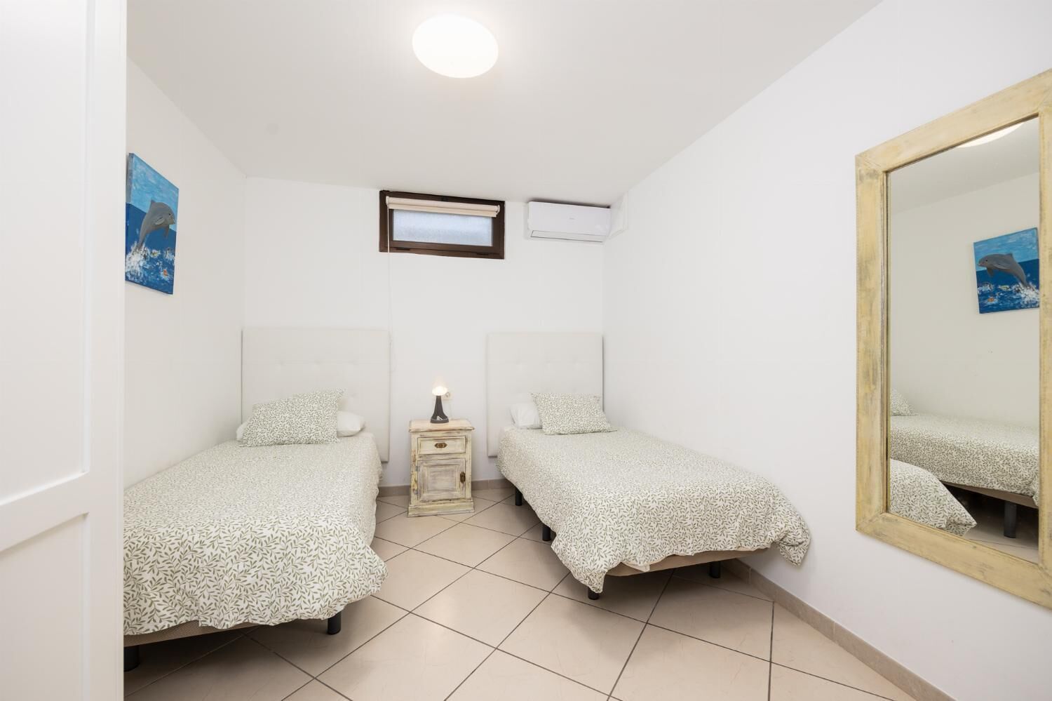 A small, clean room with two single beds, a nightstand with a lamp, a window with blinds, and a ceiling light.