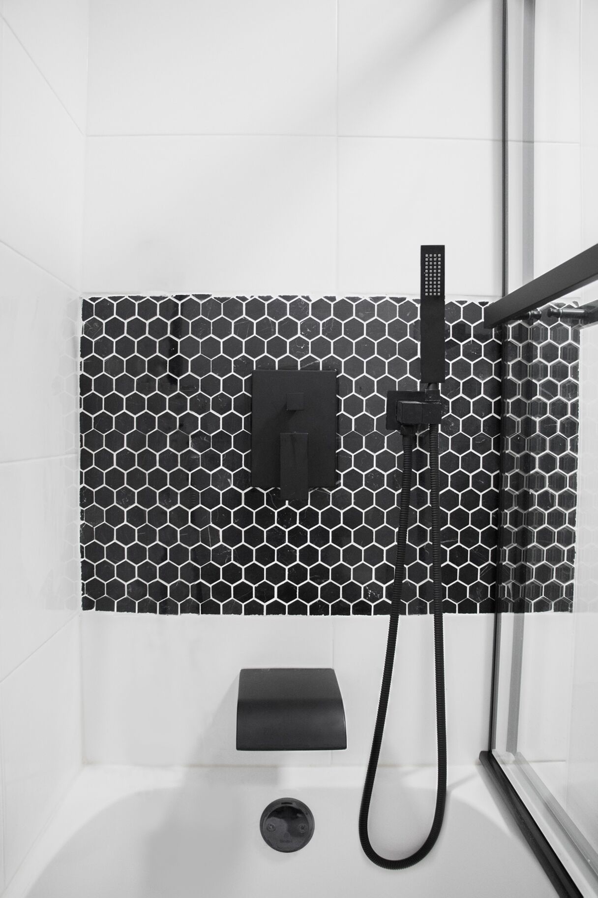 Un espacio de ducha moderno con un resguardo de azulejos hexagonales blanco y negro, un cabezal de ducha de mano y un panel de control.