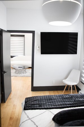 Een moderne slaapkamer met een bed, een stoel, een aan de muur bevestigde televisie en een deur naar een andere kamer.