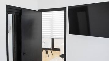 Une chambre moderne avec un lit, un bureau et une chaise. Une porte donne accès à une autre pièce, et il y a une fenêtre avec des stores.