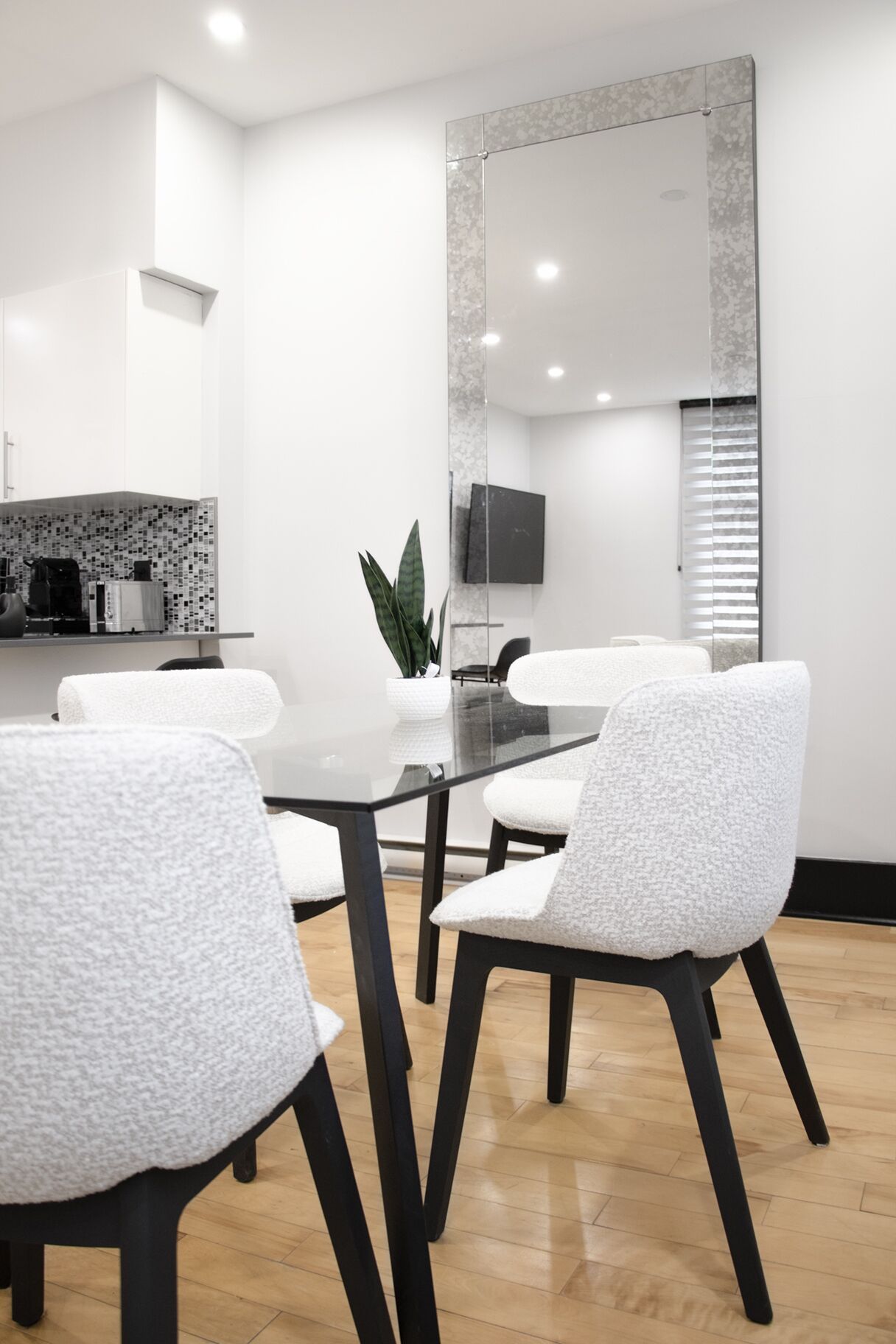 A modern dining area with a glass table and white cushioned chairs, a potted plant, and a large mirror on the wall.