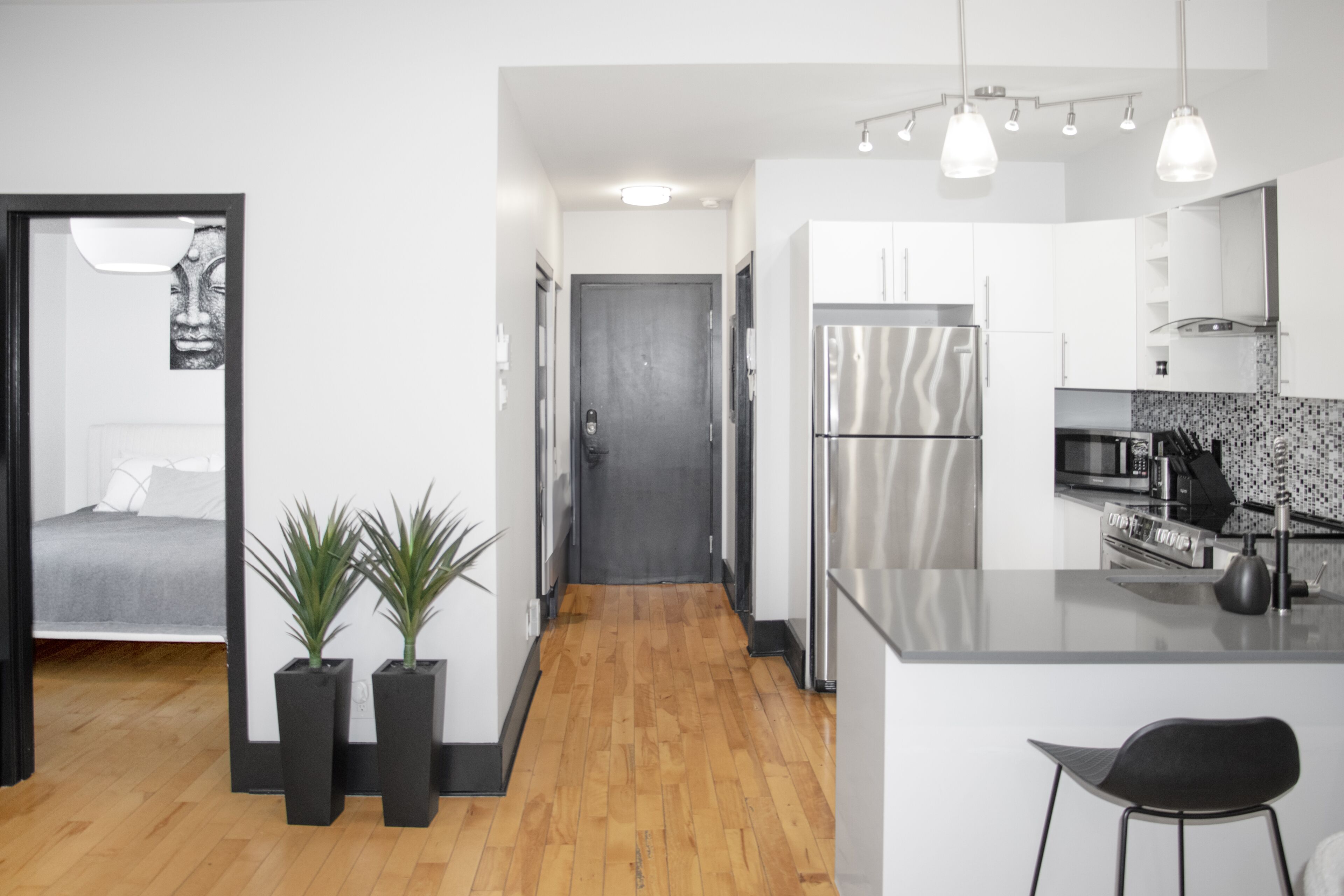 A modern kitchen with a stainless steel refrigerator, a dining area with a black chair, and a hallway leading to a bedroom with a bed and a wall-mounted artwork.