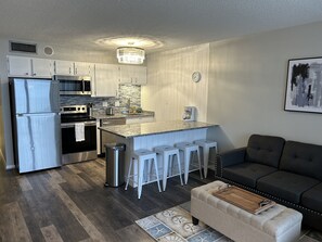 A compact kitchen with a refrigerator, stove, and sink, a dining area with stools, and a living room with a sofa.