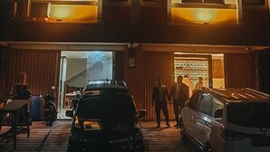 Un edificio de varios pisos con fachada de ladrillo y ventanas amplias. Hay coches estacionados adelante y personas caminando cerca de la entrada.