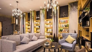 Un salon moderne avec un canapé gris, un fauteuil bleu et une table basse ronde. De hautes étagères remplies de livres et d’objets décoratifs ornent la pièce.